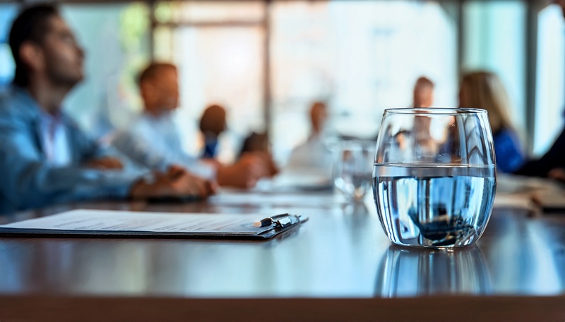 Glas Wasser auf Konferenztisch, Geschäftspartner im Hintergrund unscharf