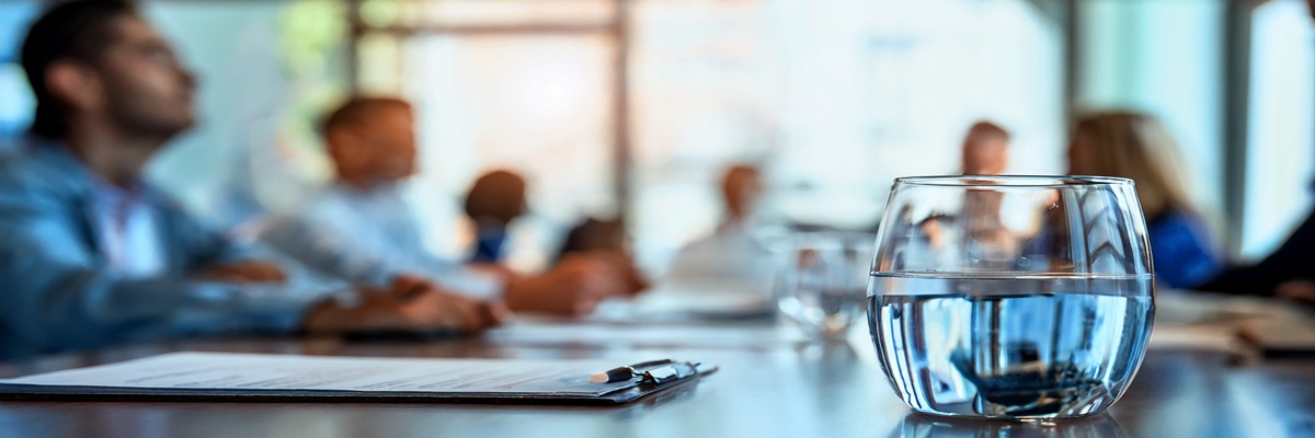 Glas Wasser auf Konferenztisch, Geschäftspartner im Hintergrund unscharf